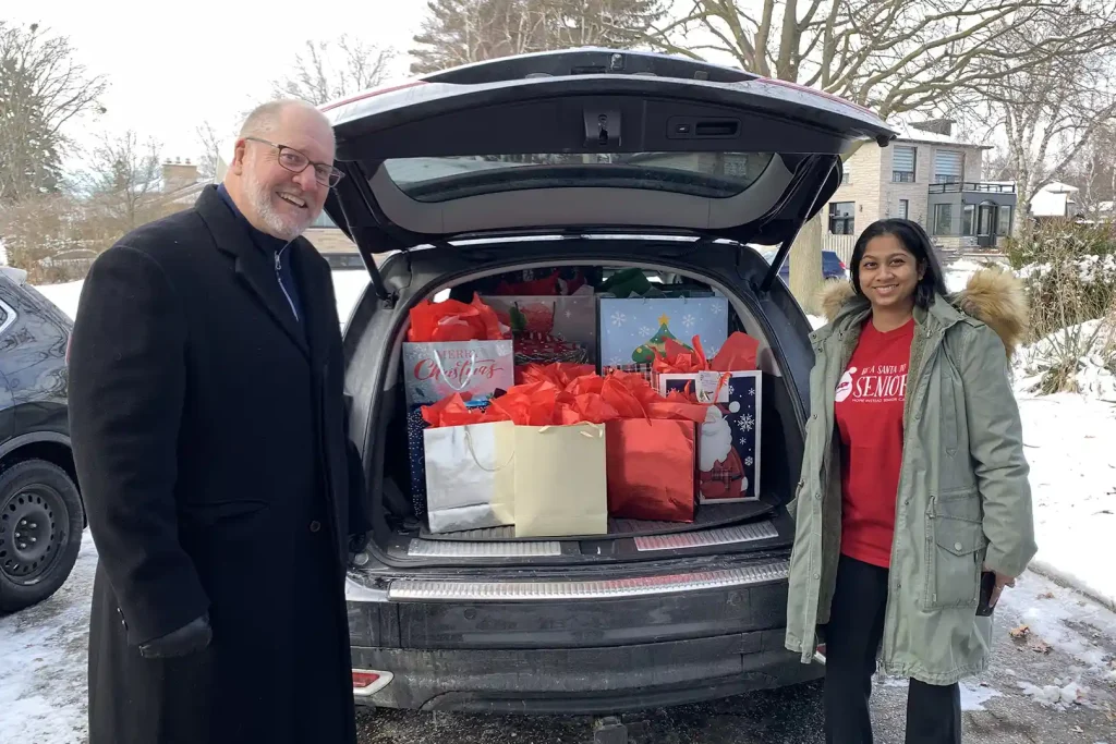 Home Instead Toronto East staff members standing beside open vehicle trunk filled with wrapped holiday gifts in red bags for Be A Santa To A Senior program