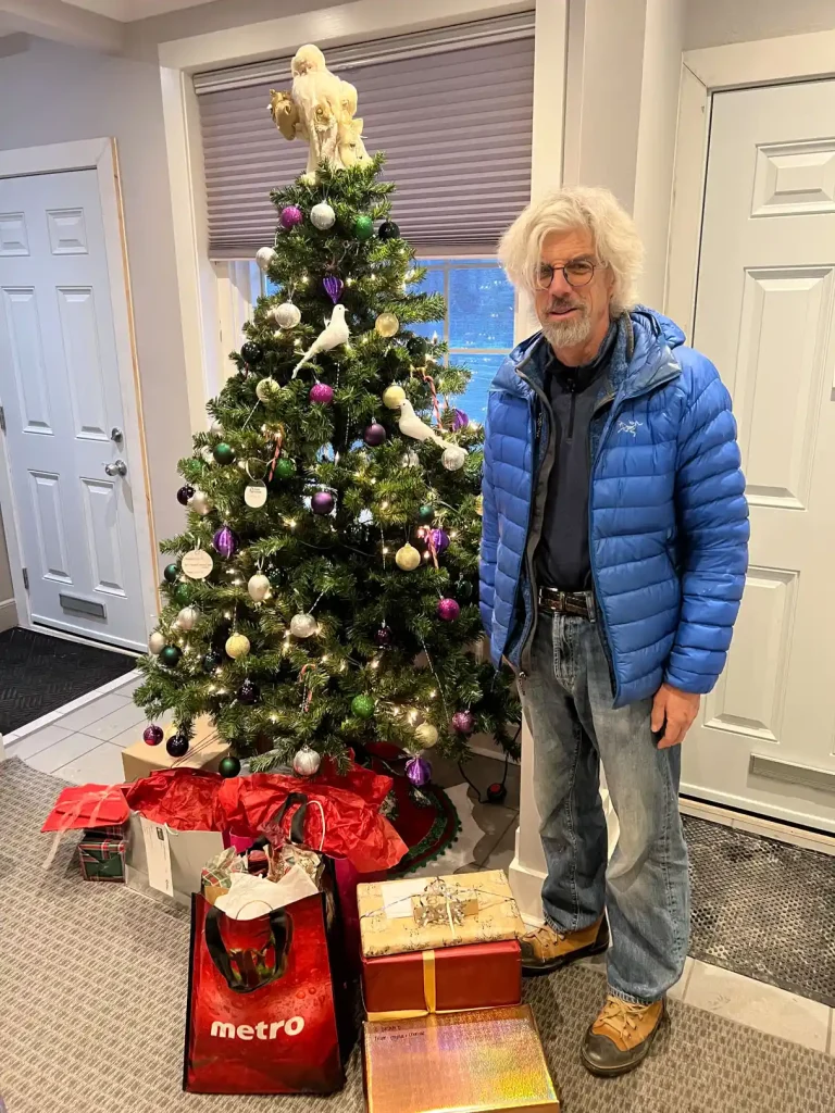 Community sponsor standing beside decorated Christmas tree with wrapped gifts and Metro shopping bag at Home Instead Toronto East office entrance