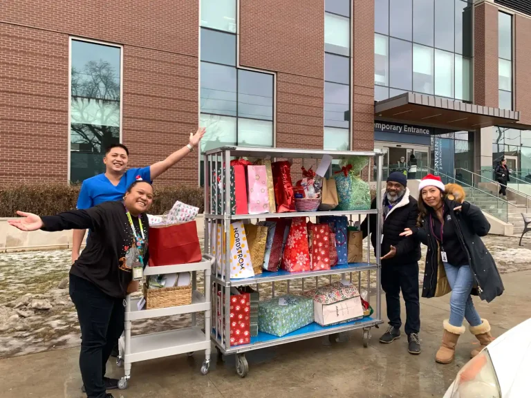Home Instead Toronto East staff and Michael Garron Hospital team members celebrating beside rolling cart loaded with colorful wrapped holiday gifts outside hospital entrance