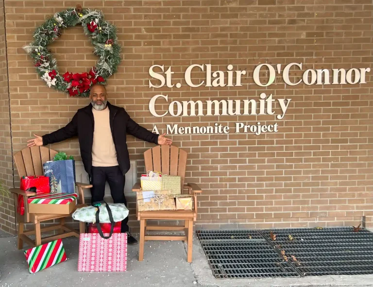 Allan Skeete, Community Relations Manager, standing with wrapped holiday gifts displayed on wooden chairs outside St. Clair O'Connor Community entrance with festive wreath