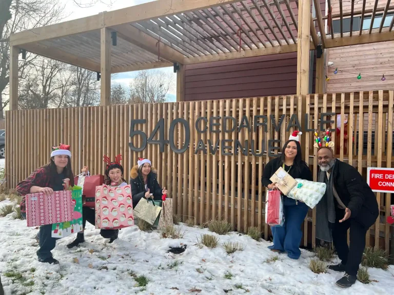 Home Instead Toronto East team wearing festive holiday accessories standing with wrapped gifts outside 540 Cedar Park Avenue Woodgreen Community Services entrance in winter