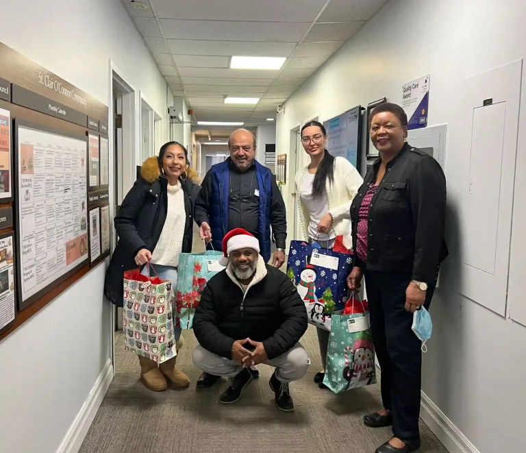Home Instead Toronto East team and St. Clair O'Connor Community staff posing together in facility hallway holding festive gift bags for senior residents