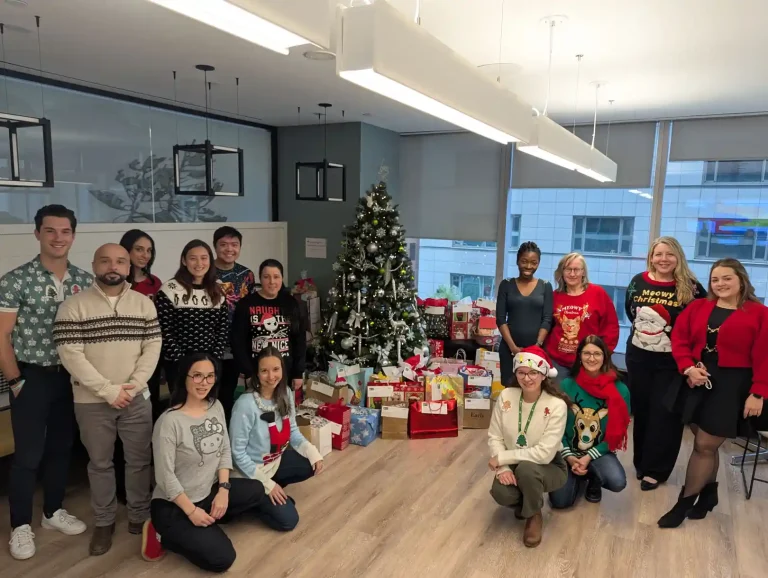 Trisura Insurance team wearing festive holiday sweaters gathered around decorated Christmas tree with wrapped gifts for Be A Santa To A Senior program