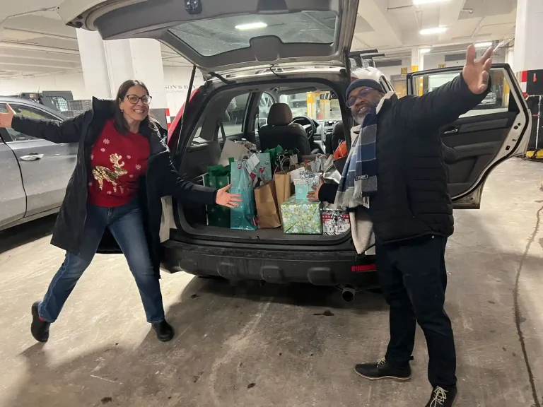 Home Instead Toronto East team members celebrating beside vehicle filled with wrapped holiday gifts and festive gift bags in parking garage