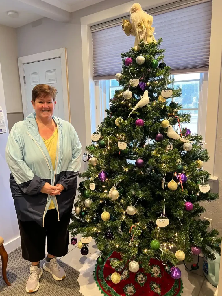 Community donor Tracy Miller standing beside decorated Christmas tree with gift tag ornaments at Home Instead Toronto East office entrance